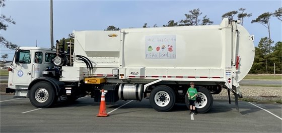 A trash truck featuring a child's artwork on the side with the artist standing in front of it